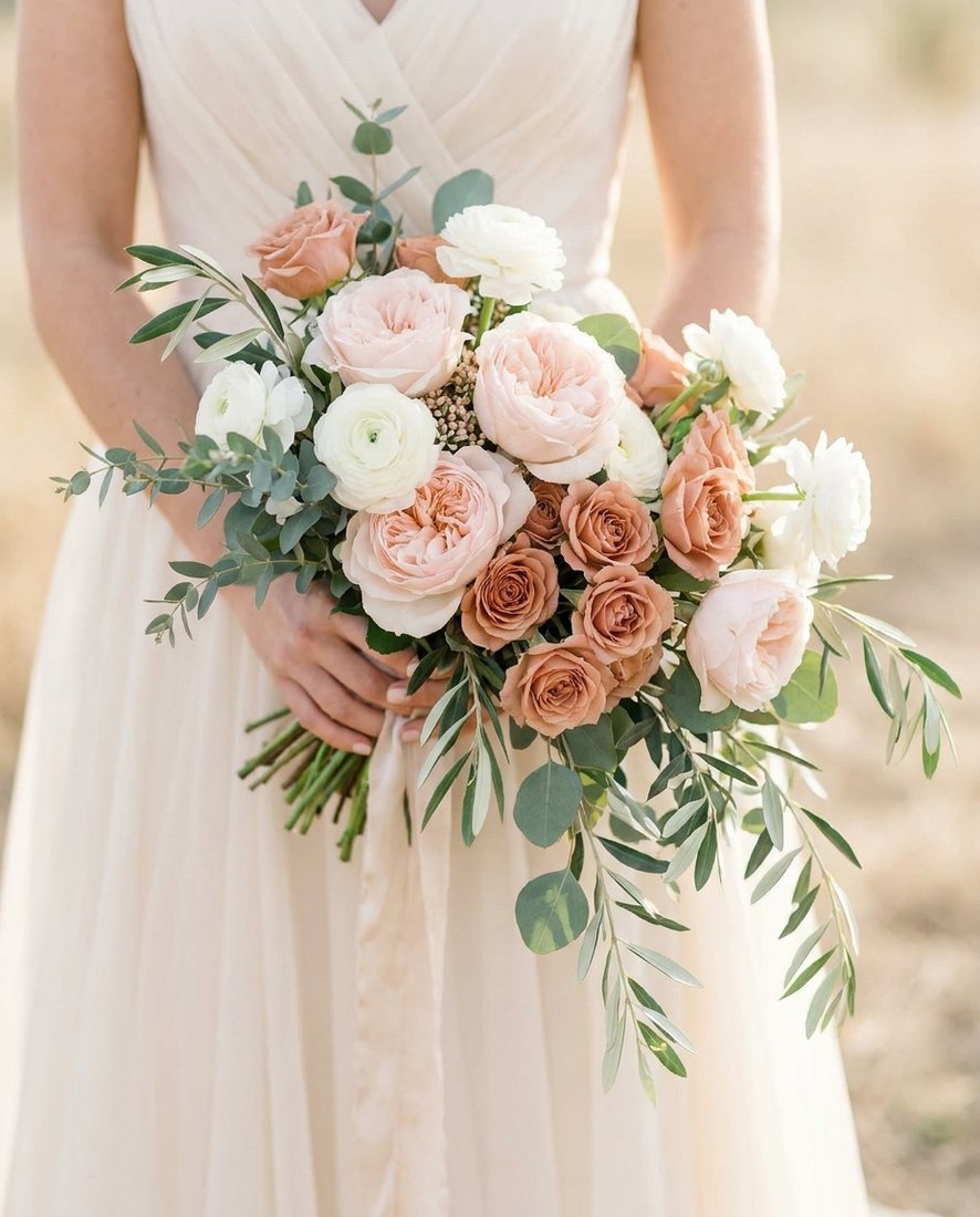 Bruidsboeket van zachte witte en blush rozen met ranonkels, vastgehouden tegen een crème trouwjurk